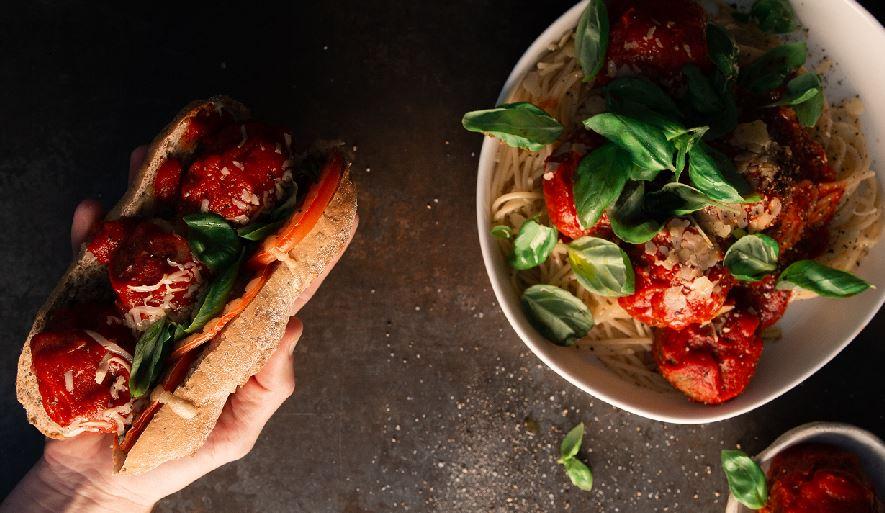Image of hand holding a meatball sub and bowl of pasta with meatballs